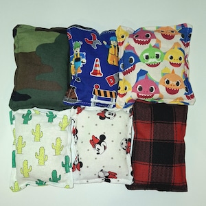 Peut inclure: Six sacs de haricots en tissu avec différents motifs. Les sacs de haricots sont tous carrés et ont des couleurs et des motifs différents. Les motifs comprennent le camouflage, les ouvriers du bâtiment, le requin bébé, le cactus, Mickey Mouse et le plaid.