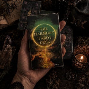 Puede incluir: Una caja rectangular de cartas de tarot titulada The Harmony Tarot Deck. La caja presenta un paisaje oscuro y atmosf&eacute;rico con un anillo dorado brillante en el centro. El texto incluye Leedesign Studio Presents y Designed by Christopher Lee.