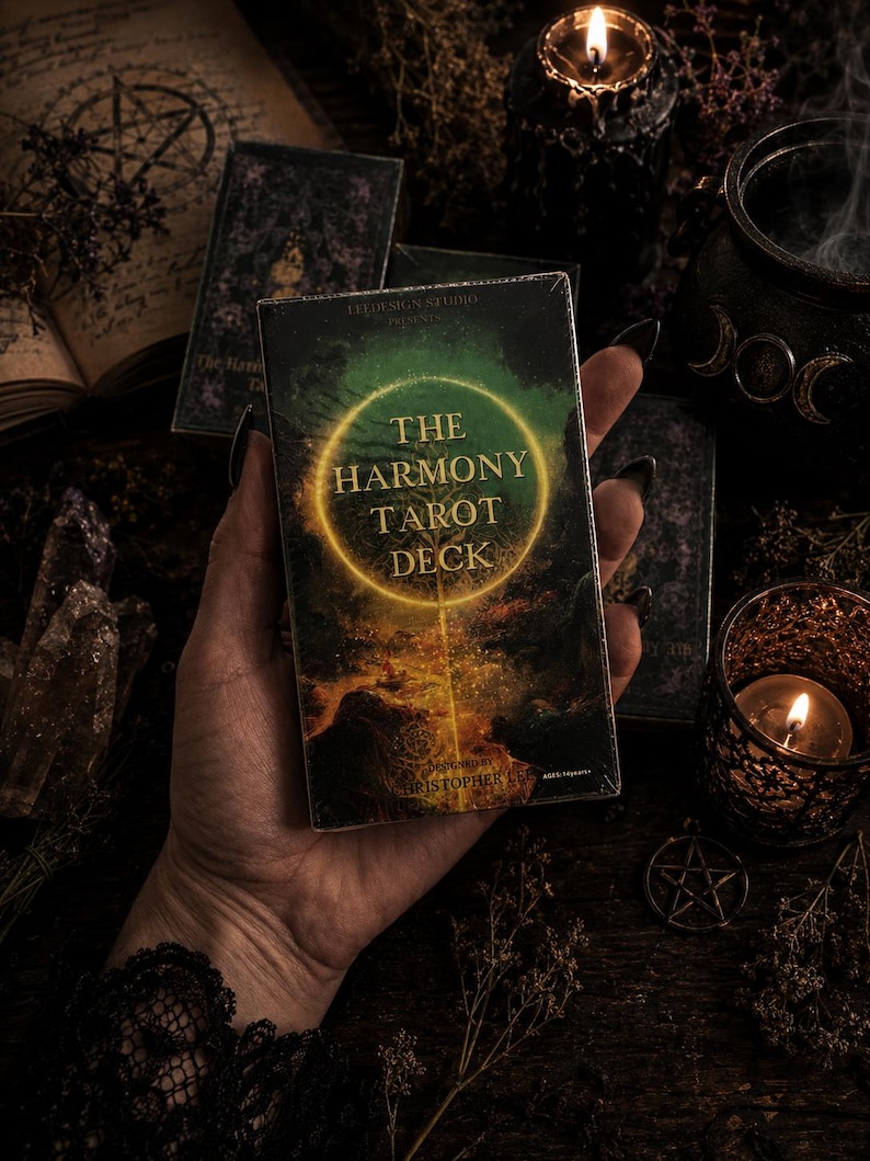 Puede incluir: Una caja rectangular de cartas de tarot titulada The Harmony Tarot Deck. La caja presenta un paisaje oscuro y atmosf&eacute;rico con un anillo dorado brillante en el centro. El texto incluye Leedesign Studio Presents y Designed by Christopher Lee.