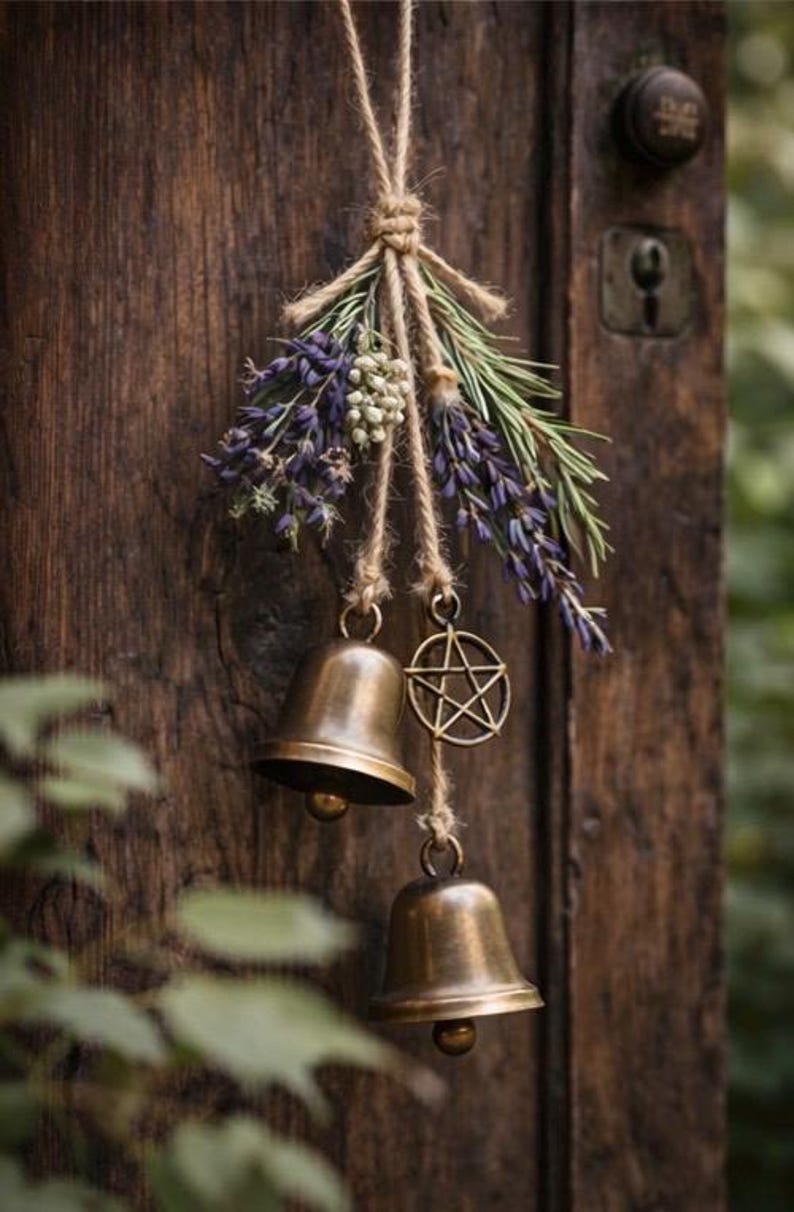 May include: A rustic door decoration featuring two bronze bells, a pentagram charm, and sprigs of lavender and greenery, all suspended by twine. The wooden door has a dark, aged appearance.