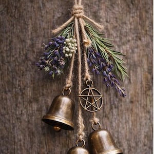 May include: A rustic hanging decoration featuring three bronze bells, a pentagram charm, and sprigs of lavender and greenery. The arrangement is suspended from a natural twine cord against a weathered wood background.
