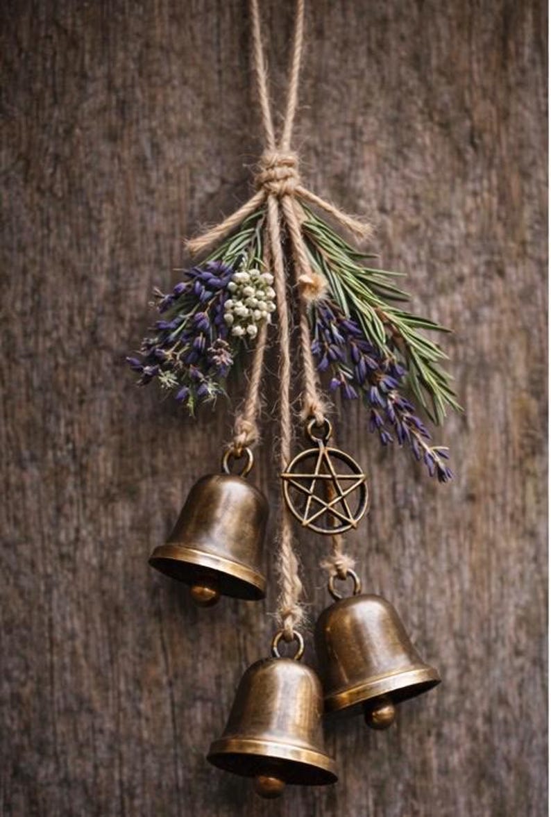 May include: A rustic hanging decoration featuring three bronze bells, a pentagram charm, and sprigs of lavender and greenery. The arrangement is suspended from a natural twine cord against a weathered wood background.