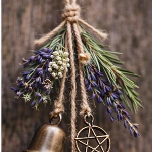 May include: A rustic hanging decoration featuring a bronze bell and a pentagram charm suspended from twine. The arrangement includes sprigs of lavender, white flowers, and green foliage, creating a natural, earthy aesthetic.