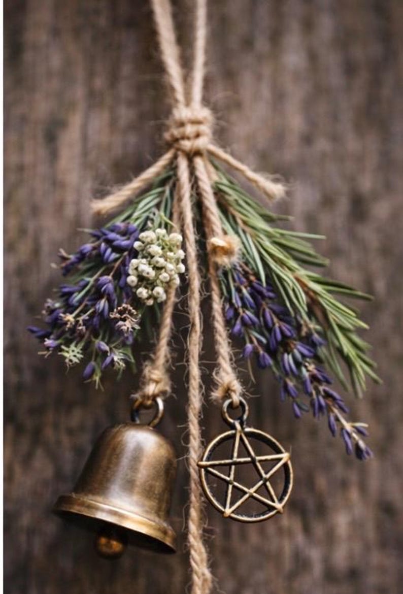 May include: A rustic hanging decoration featuring a bronze bell and a pentagram charm suspended from twine. The arrangement includes sprigs of lavender, white flowers, and green foliage, creating a natural, earthy aesthetic.