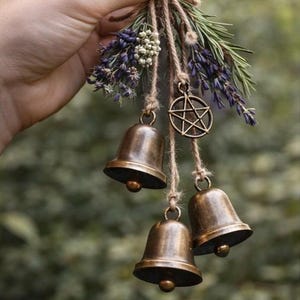 May include: A rustic hanging decoration featuring three bronze bells suspended from twine, adorned with sprigs of lavender, greenery, and a bronze pentagram charm. The bells have a warm, aged finish.