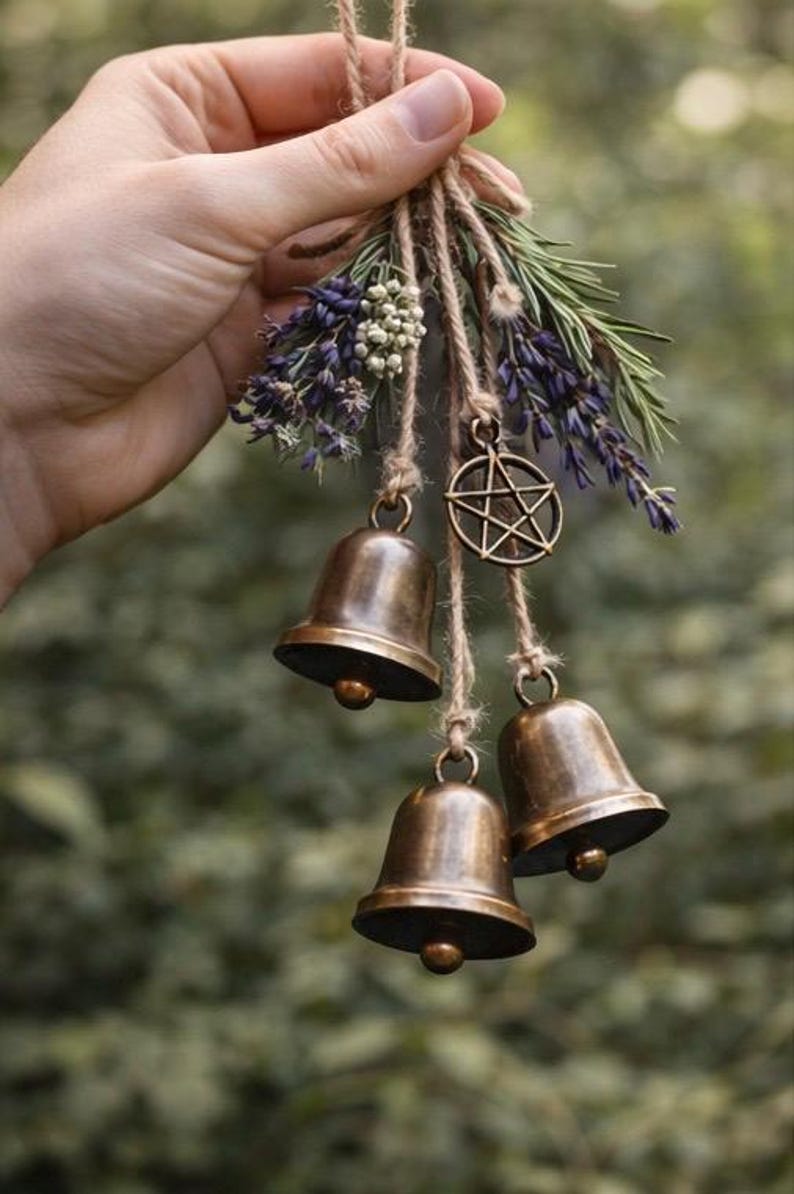 May include: A rustic hanging decoration featuring three bronze bells suspended from twine, adorned with sprigs of lavender, greenery, and a bronze pentagram charm. The bells have a warm, aged finish.