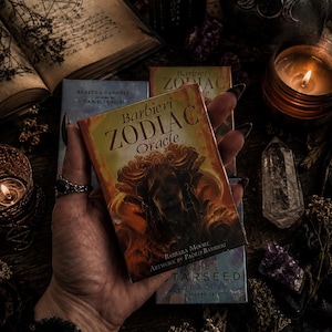 Puede incluir: Una baraja rectangular de cartas Barbieri Zodiac Oracle con una ilustraci&oacute;n de color naranja y dorado de una figura en la portada, con el t&iacute;tulo Barbieri ZODIAC Oracle y los nombres Barbara Moore y Paolo Barbieri impresos en ella.