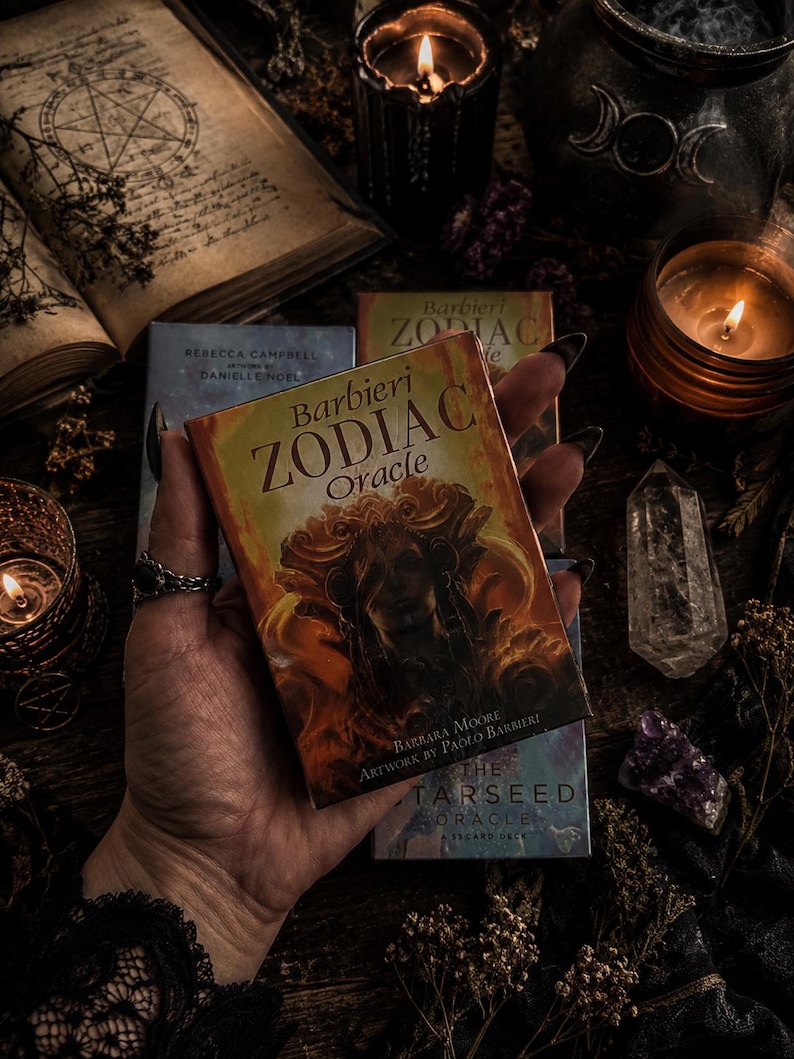 Puede incluir: Una baraja rectangular de cartas Barbieri Zodiac Oracle con una ilustraci&oacute;n de color naranja y dorado de una figura en la portada, con el t&iacute;tulo Barbieri ZODIAC Oracle y los nombres Barbara Moore y Paolo Barbieri impresos en ella.