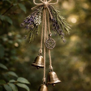 May include: A rustic wind chime featuring three bronze bells suspended from twine. The chime is adorned with dried lavender, greenery, and a metal pentacle charm. The arrangement is tied with a natural twine bow, creating a natural, earthy aesthetic.