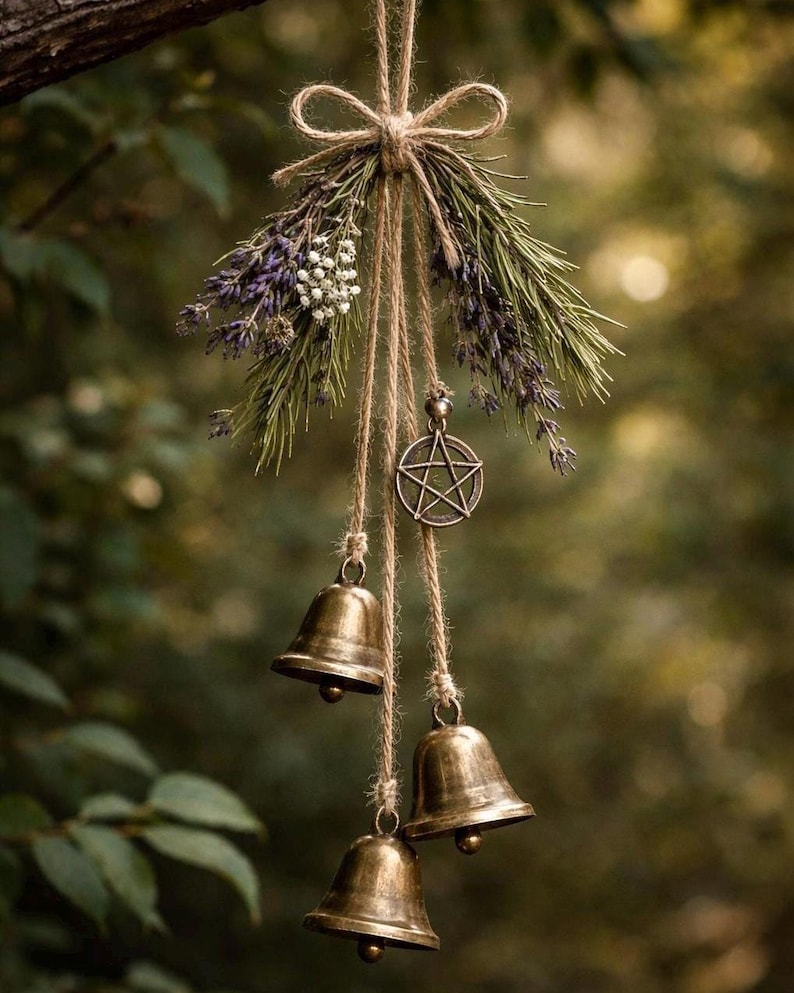 May include: A rustic wind chime featuring three bronze bells suspended from twine. The chime is adorned with dried lavender, greenery, and a metal pentacle charm. The arrangement is tied with a natural twine bow, creating a natural, earthy aesthetic.