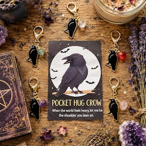 May include: A collection of crow-themed keychains with various gemstone accents, displayed with a candle, dried lavender, and a card that reads "Pocket Hug Crow". The keychains feature a black crow design.