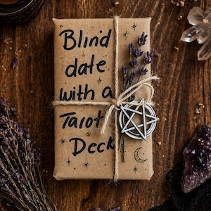 Puede incluir: Un paquete rectangular envuelto en papel marr&oacute;n con el texto Blind date with a Tarot Deck escrito en tinta negra. El paquete est&aacute; atado con cordel y decorado con una ramita de lavanda seca y un dije de pentagrama de metal plateado.