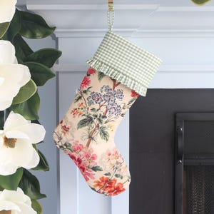 May include: A Christmas stocking with a floral pattern in pink, red, and blue on a cream background. The top is green and white gingham with a ruffle. A gold-colored metal hook holds the stocking.