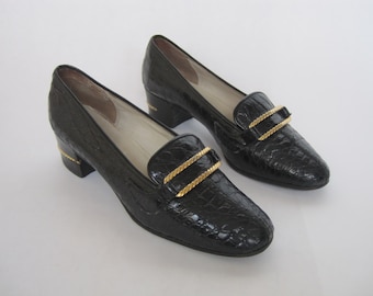 Könnte beinhalten: Paar schwarze Leder-Schnallen-Slipper mit goldenen Schnallen. Die Slipper haben ein Kroko-Prägemuster.
