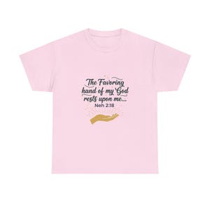 May include: A light pink t-shirt with the text "The Favoring hand of my God rests upon me... Neh 2:18" in black script. Below the text is a gold-colored hand design with radiating lines.