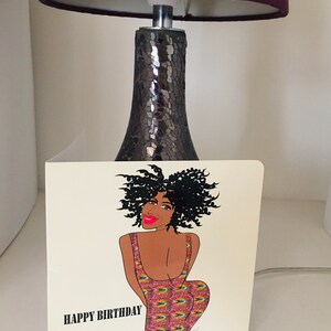 May include: A birthday card featuring a cartoon illustration of a woman with curly black hair wearing a colorful patterned dress. The card has the text "HAPPY BIRTHDAY" printed on the bottom left corner.