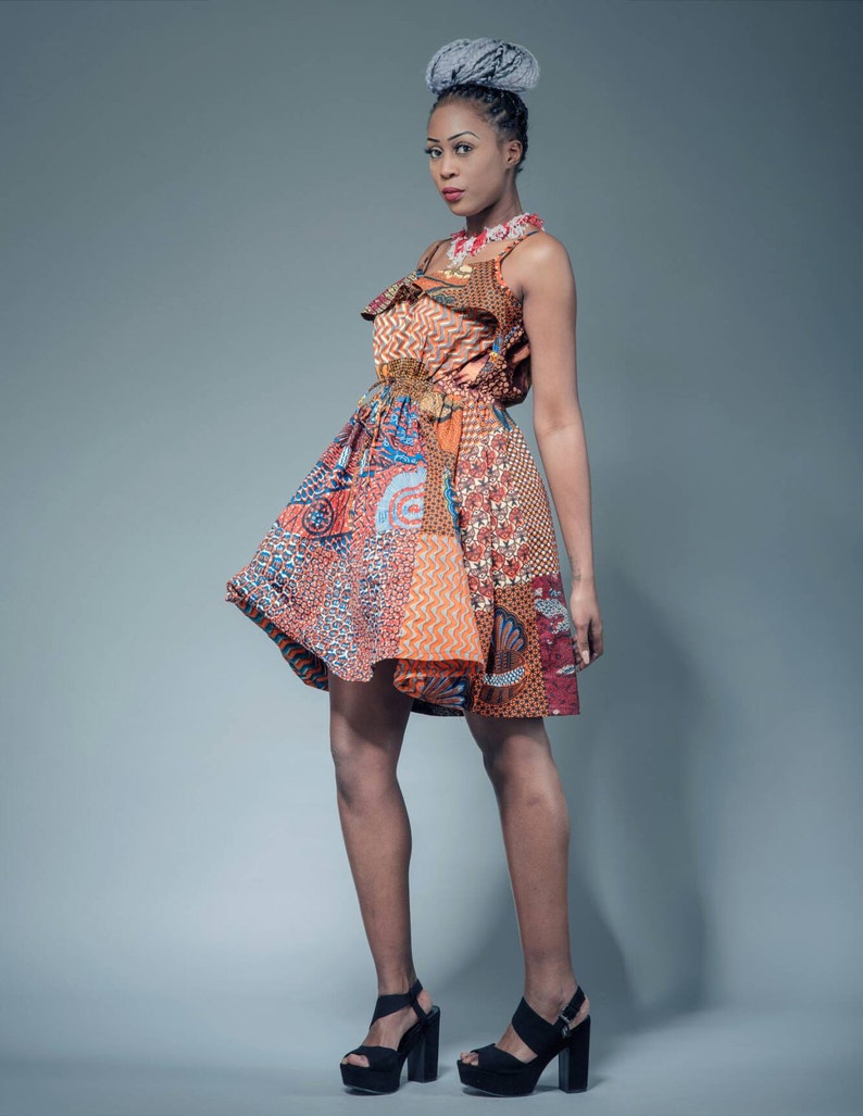 May include: A woman wearing a colorful patterned dress with a ruffled neckline and a gray hair bun. She is wearing black heeled sandals and has a necklace on.