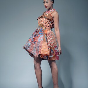 May include: A woman wearing a colorful, patterned dress with a ruffled neckline and a belt. She has gray hair in a bun and is wearing black heeled sandals.