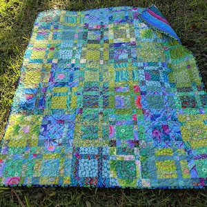Puede incluir: Una colcha de retazos con una combinación de colores azules y verdes. La colcha está hecha de muchos cuadrados de tela diferentes, cada uno con un patrón único. La colcha está ribeteada con un borde negro y blanco.