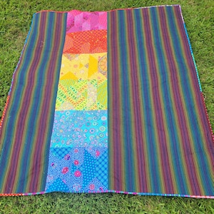 May include: A colorful quilt with a central panel of patchwork in pink, orange, yellow, green, turquoise, and blue. The sides feature vertical stripes in rainbow hues. The quilt is laid out on green grass.