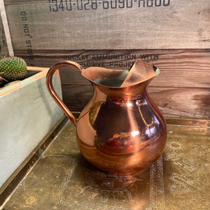 May include: A copper pitcher with a handle and a shiny, reflective surface. The pitcher is sitting on a brass tray with an ornate design.