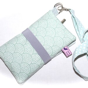 May include: A light green phone case with a white polka dot pattern and a gray strap. The case has a small pink label that says "S&uuml;&szlig;e Handarbeit". The case is attached to a light green lanyard with white stars.