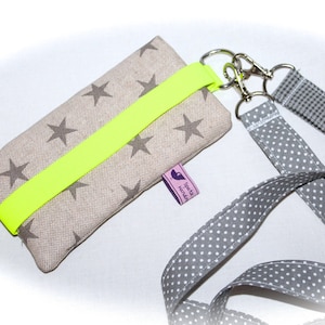 Puede incluir: Una funda de tel&eacute;fono beige con una correa amarilla ne&oacute;n y un cord&oacute;n gris y blanco con lunares. La funda tiene un patr&oacute;n de estrellas y una peque&ntilde;a etiqueta morada que dice "Spot It Handmade".