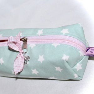 Op de afbeelding: Een lichtgroene ritszak met een wit sterrenpatroon. De tas heeft een roze rits en een roze-witte gingham strik met een zilveren hanger.