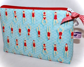 Peut inclure: Une pochette zippée en tissu bleu et blanc avec un ruban en vichy rouge et blanc et un petit charme. La pochette présente un motif répété de figures rouges en maillot de bain nageant dans un motif de vagues bleu et blanc. La pochette a une étiquette violette avec le texte "Sea Salt Handmade".