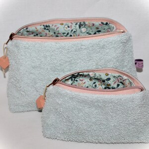 Może przedstawiać: Dwie jasnoniebieskie kosmetyczki zapinane na zamek błyskawiczny z różową lamówką i podszewką z kwiatowym wzorem. Większa kosmetyczka znajduje się na mniejszej.