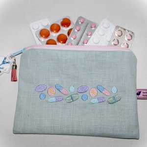 Könnte beinhalten: Eine hellblaue, gepolsterte Kosmetiktasche aus Leinen mit einem rosa Reißverschluss und einem rosa Quastenanhänger. Die Tasche hat ein gesticktes Design aus bunten Pillen und Kapseln. An dem Reißverschluss ist ein kleiner weißer Arztkittel-Anhänger befestigt. Die Tasche hat ein lilafarbiges Etikett mit dem Text "Spotta Handmade".