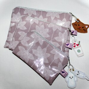 Könnte beinhalten: Drei Reißverschlusstaschen mit einem Blumenmuster in Grautönen und Weiß. Die Taschen sind mit kleinen weißen Punkten verziert. Die Taschen haben Reißverschluss-Zipper mit Anhängern, darunter eine Katze, eine Kaffeetasse und eine Menstruationstasse.