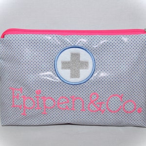 Könnte beinhalten: Eine weiße Reißverschlusstasche mit einem rosa Reißverschluss und Besatz. Die Tasche hat ein blaues und weißes Punkte-Muster und zeigt ein weiß gesticktes Kreuz in einem blauen Kreis. Der Text "EpiPen & Co." ist in rosa auf der Vorderseite der Tasche gestickt. Ein kleiner Schlüsselanhänger mit einem roten und gelben EpiPen ist am Reißverschluss befestigt.