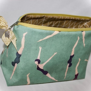 Puede incluir: Un neceser turquesa con un estampado repetido de buceadores con trajes de baño negros. El neceser tiene una cremallera amarilla y un lazo de cuadros. El forro interior es un estampado geométrico en marrón y blanco.