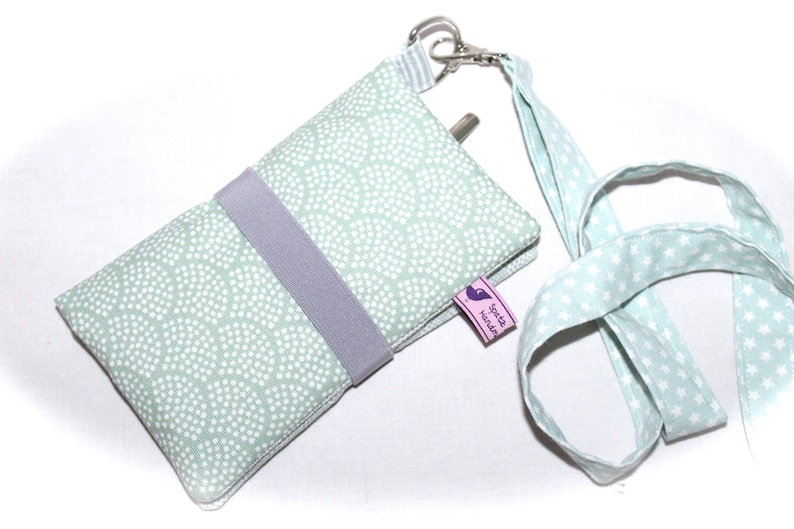 May include: A light green phone case with a white polka dot pattern and a gray strap. The case has a small pink label that says "S&uuml;&szlig;e Handarbeit". The case is attached to a light green lanyard with white stars.