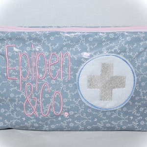 Könnte beinhalten: Eine hellblaue Reißverschlusstasche mit einem weißen Kreuz auf einem blauen Kreis und dem gestickten Text "Epipen & Co." in Rosa. Die Tasche hat einen kleinen weißen und blauen Arzt-Anhänger, der am Reißverschluss befestigt ist.