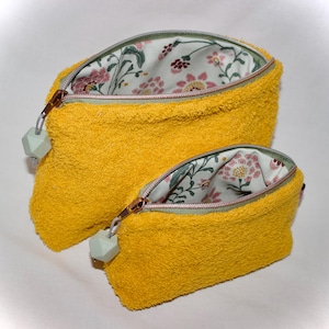 Może przedstawiać: Dwie żółte kosmetyczki zapinane na zamek błyskawiczny z podszewką z tkaniny w kwiatowy wzór. Większa kosmetyczka ma jasnozielony suwak z jasnozielonym geometrycznym brelokiem. Mniejsza kosmetyczka ma jasnozielony suwak z jasnozielonym geometrycznym brelokiem.