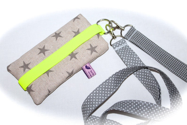 Puede incluir: Una funda de tel&eacute;fono beige con una correa amarilla ne&oacute;n y un cord&oacute;n gris y blanco con lunares. La funda tiene un patr&oacute;n de estrellas y una peque&ntilde;a etiqueta morada que dice "Spot It Handmade".