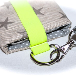 Puede incluir: Una peque&ntilde;a bolsa de tela beige con una correa amarilla ne&oacute;n y un cierre de metal plateado. La bolsa tiene un forro de lunares blancos y grises y un patr&oacute;n de estrellas grises y blancas en el exterior.