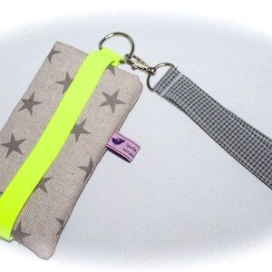Puede incluir: Una funda para tel&eacute;fono de tela beige con una correa amarilla ne&oacute;n y una peque&ntilde;a etiqueta morada que dice "spatz hundin". La funda tiene un fondo blanco con estrellas grises impresas. La funda tiene una correa a cuadros gris y blanca unida a un llavero plateado.