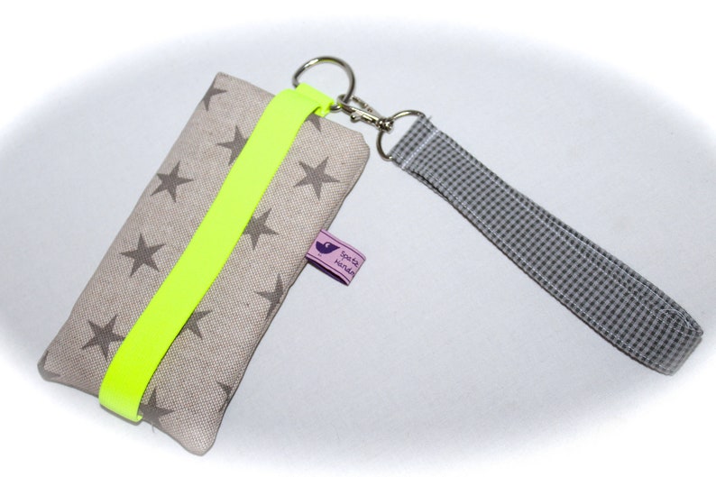 Puede incluir: Una funda para tel&eacute;fono de tela beige con una correa amarilla ne&oacute;n y una peque&ntilde;a etiqueta morada que dice "spatz hundin". La funda tiene un fondo blanco con estrellas grises impresas. La funda tiene una correa a cuadros gris y blanca unida a un llavero plateado.
