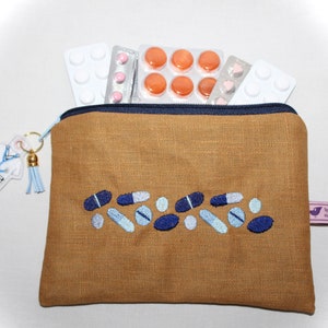 Könnte beinhalten: Eine braune Leinen-Reißverschlusstasche mit einem blauen Reißverschluss und einem blauen und weißen gestickten Design von Pillen. Die Tasche hat einen kleinen Schlüsselanhänger in Form eines Arztkittels mit einem blauen Stethoskop und einem blauen Quasten.