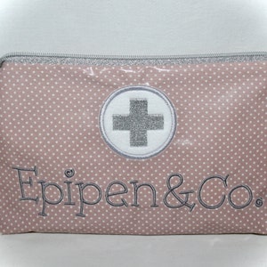 Könnte beinhalten: Eine rosa Reißverschlusstasche mit weißen Punkten. Die Tasche hat einen weißen Kreis mit einem silbernen Kreuz und den Text "Epipen & Co." in grau gestickt.