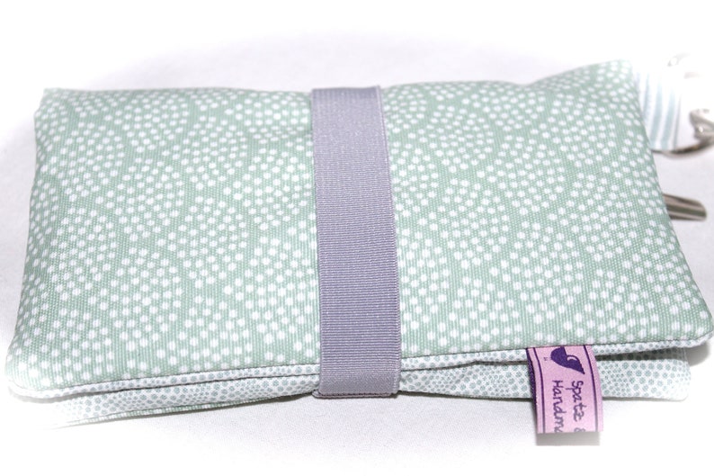 May include: A light green zippered pouch with a white polka dot pattern. The pouch has a gray fabric strap and a purple tag that says "Spatz & Handmade".