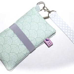 May include: A light green fabric phone case with a white polka dot pattern and a gray strap. The case has a silver key ring and a white fabric strap with white stars. The case has a small pink label with the text "Sp&auml;te N&auml;chte" in purple.