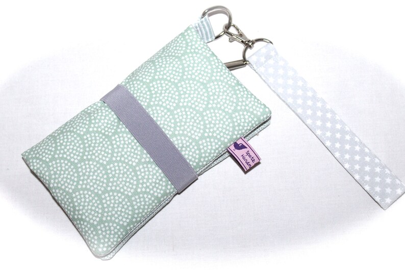 May include: A light green fabric phone case with a white polka dot pattern and a gray strap. The case has a silver key ring and a white fabric strap with white stars. The case has a small pink label with the text "Sp&auml;te N&auml;chte" in purple.