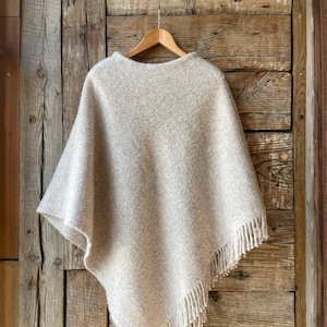 Peut inclure: Un poncho en tricot beige avec des détails en franges suspendu à un cintre en bois sur un fond en bois rustique.