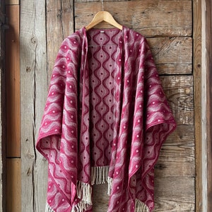 May include: A red and white patterned shawl with fringe. The shawl is hanging on a wooden hanger against a rustic wooden background.
