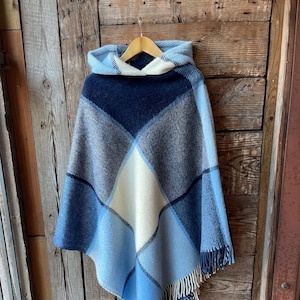 Op de afbeelding: Een blauwe en witte geruite poncho met capuchon en franjes. De poncho hangt aan een houten hanger tegen een rustieke houten achtergrond.
