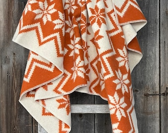 Puede incluir: Una manta de tiro de color blanco y naranja con un diseño de copo de nieve. La manta está doblada y drapeada sobre una escalera de madera.